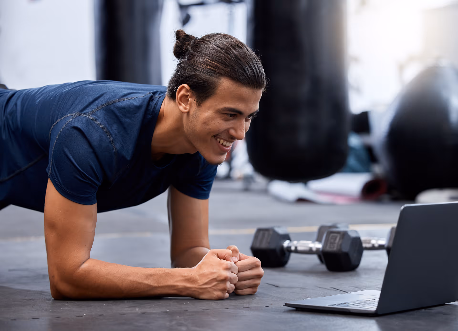 Man in a blue shirt doing a plank exercise while smiling at a laptop in a gym setting with dumbbells and punching bags in the background.