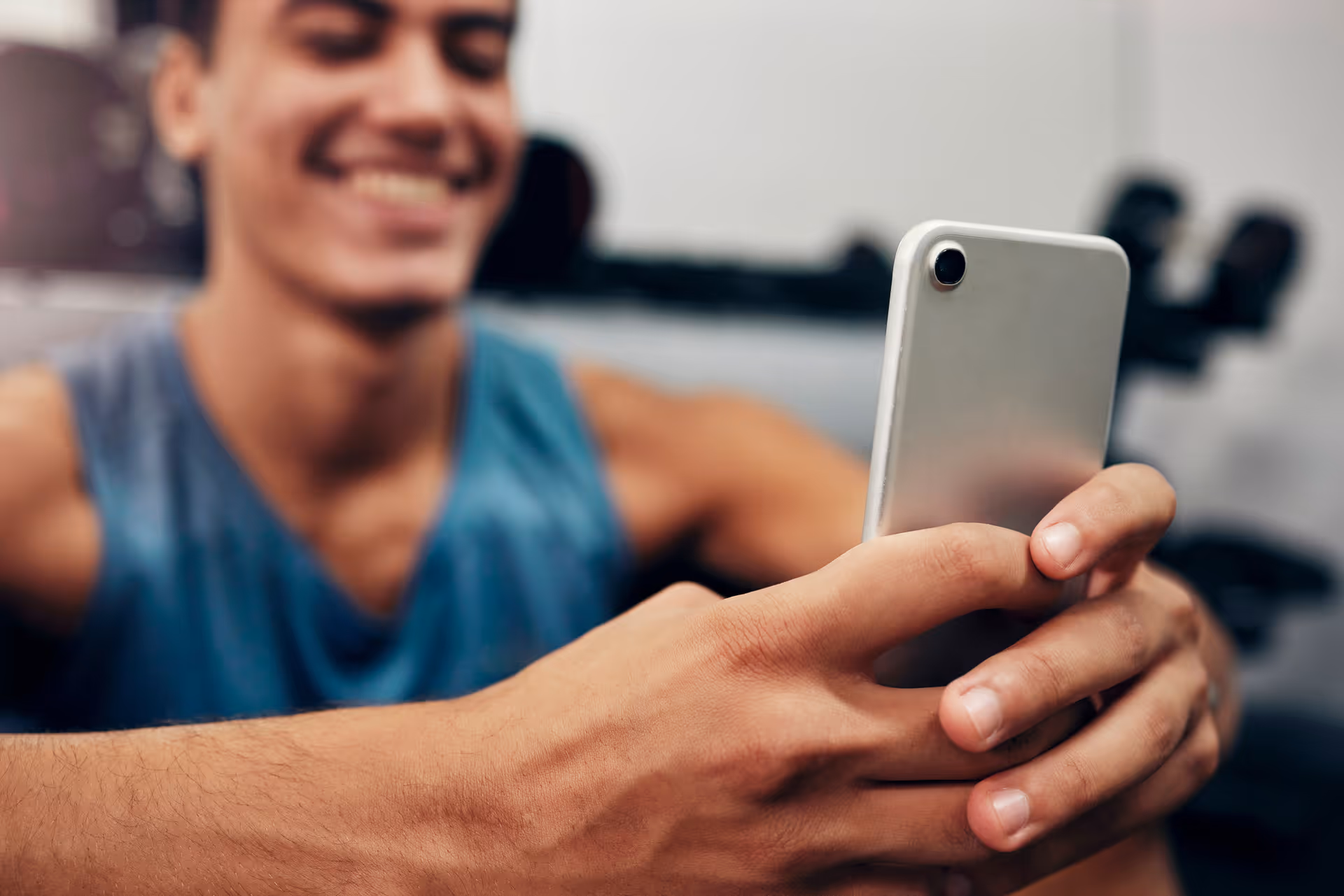 Young man in a sleeveless blue shirt smiling while holding and looking at a smartphone.