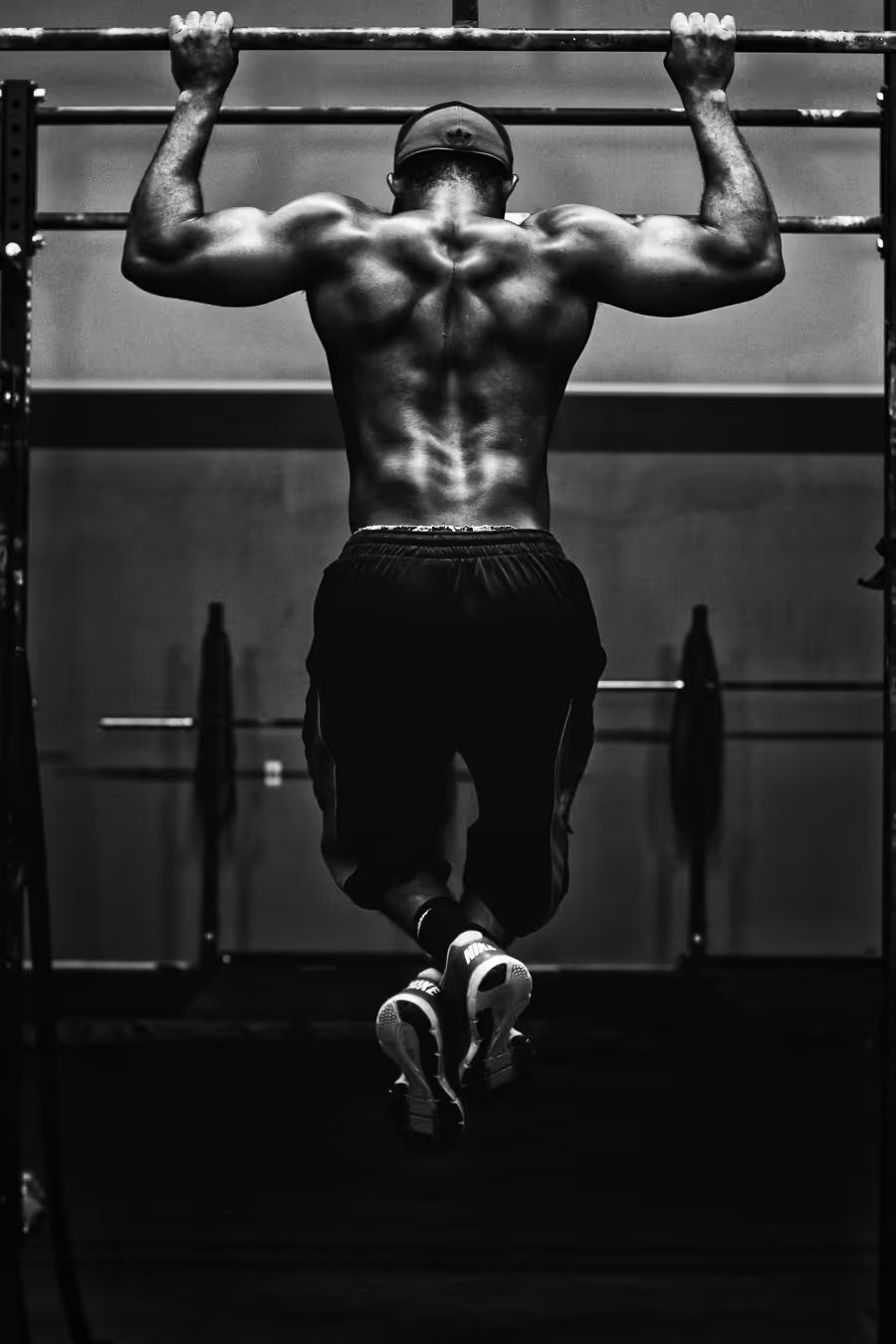 Muscular man wearing a cap performing pull-up exercise on a bar in a gym, showing defined back muscles.