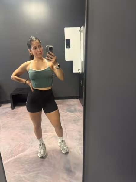 Woman in athletic wear taking a mirror selfie in a gym with a dark wall and pink floor.