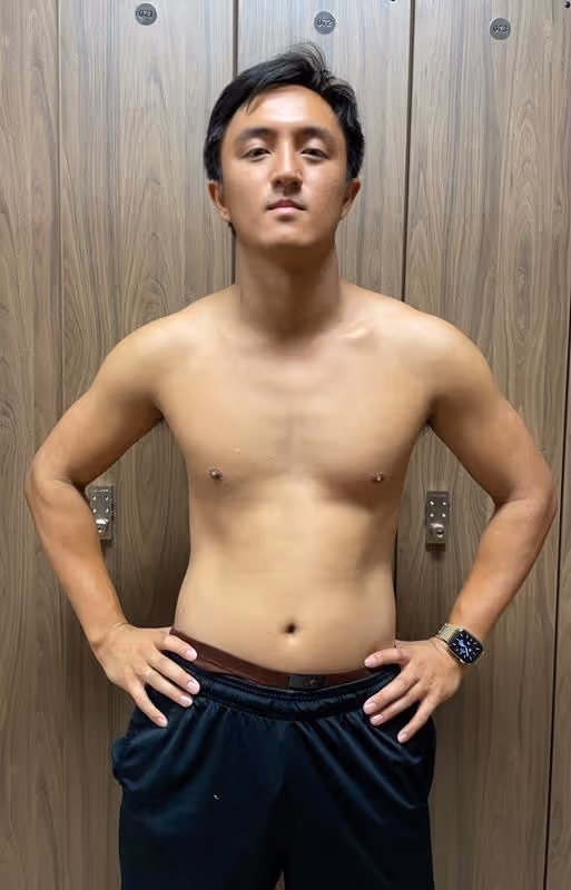 Shirtless man standing confidently with hands on hips in front of wooden lockers.