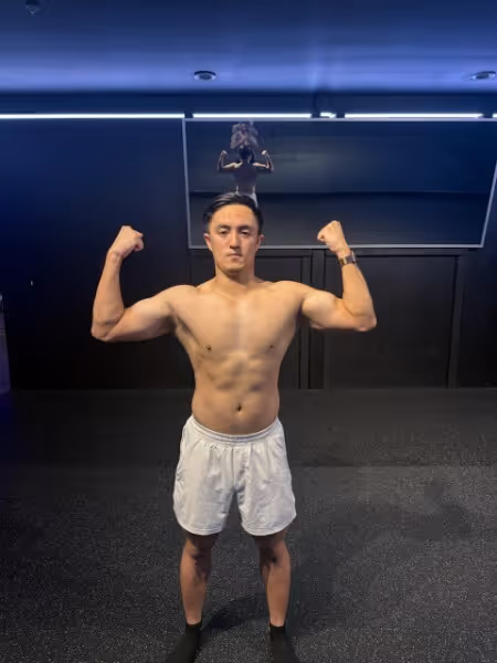 Shirtless man in white shorts flexing biceps in a gym with dark walls and ceiling lights.