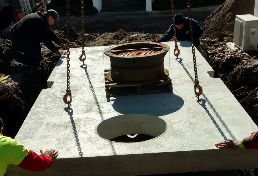 Crew guiding a concrete grease trap lid into place with chains