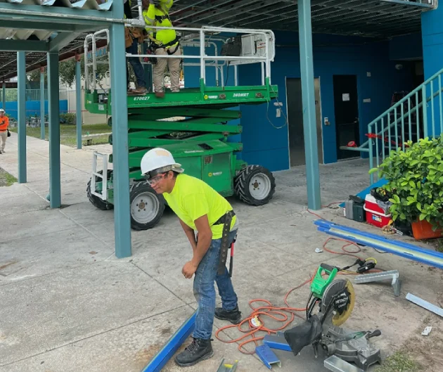 Worker cutting metal framing with a scissor lift and crew in the background