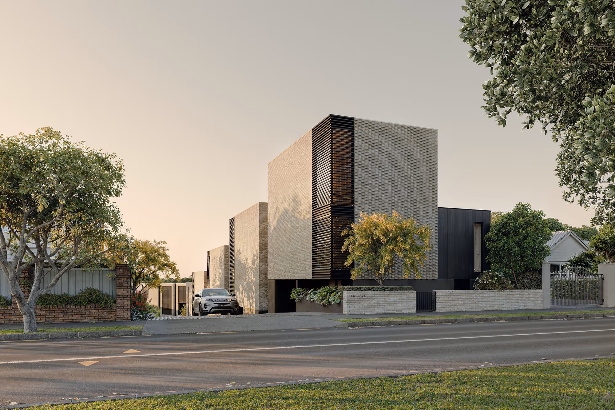 Modern residential building with cubic architecture, beige brick and black slatted accents, surrounded by trees and a car parked at the driveway.