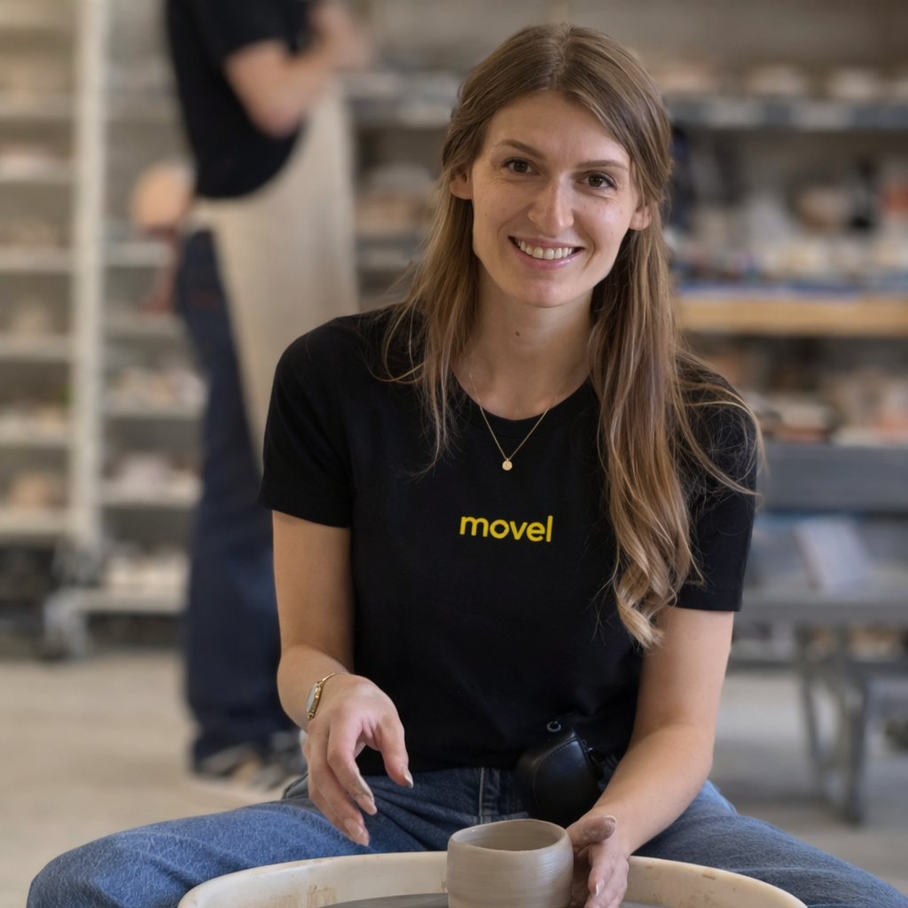 Smiling woman shaping pottery on a wheel in a studio wearing a black movel shirt.