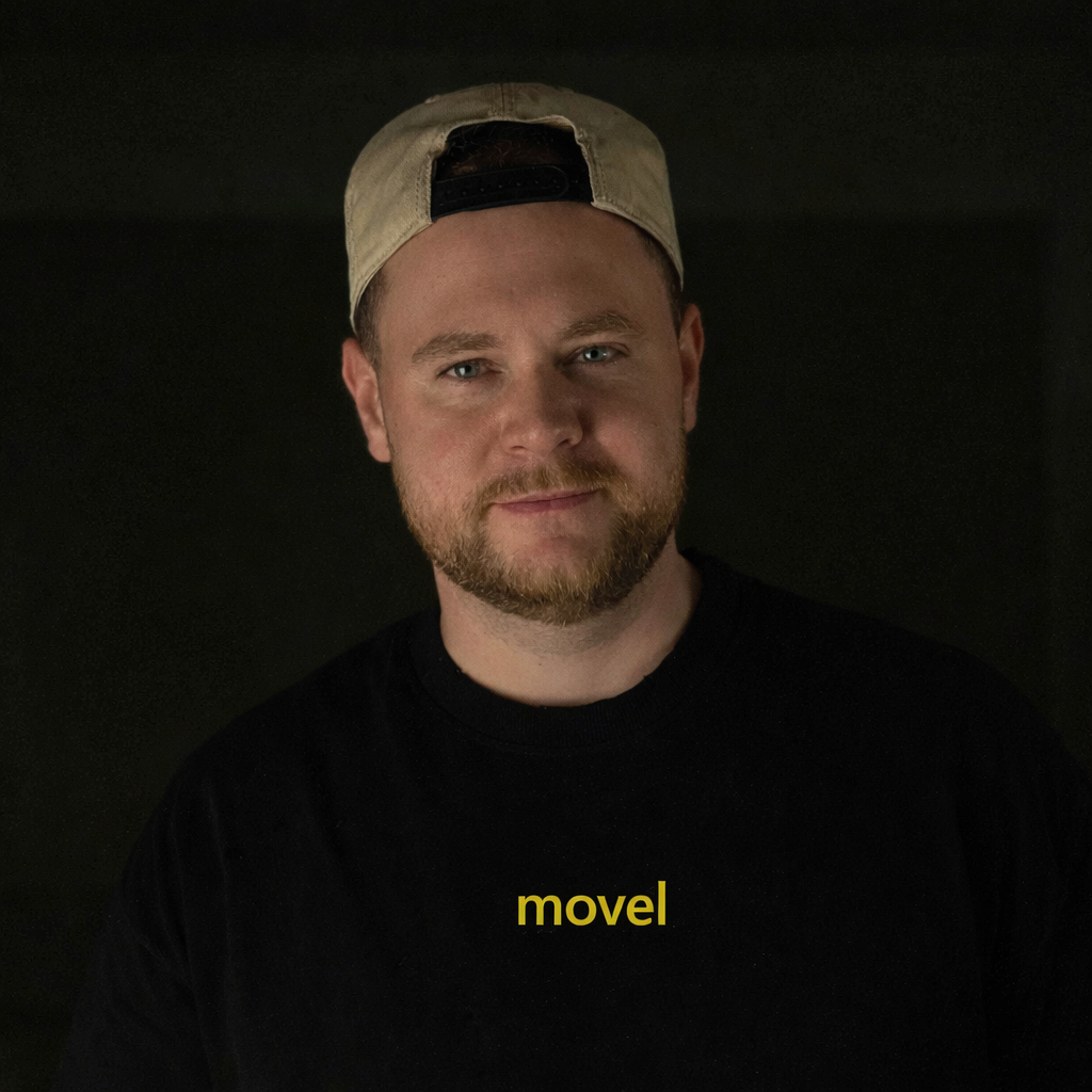 Man with beard wearing a backward beige cap and black shirt with yellow text 'movel' on a dark background.