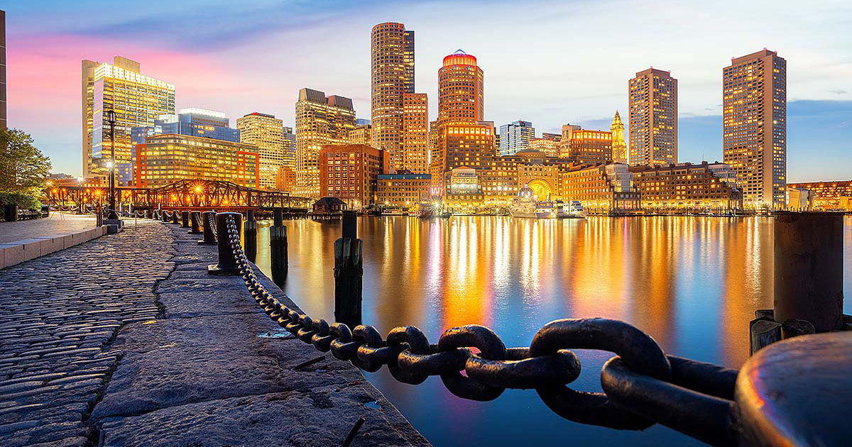Evening view of Boston harbor waterfront with city skyline and glowing building reflections on water.