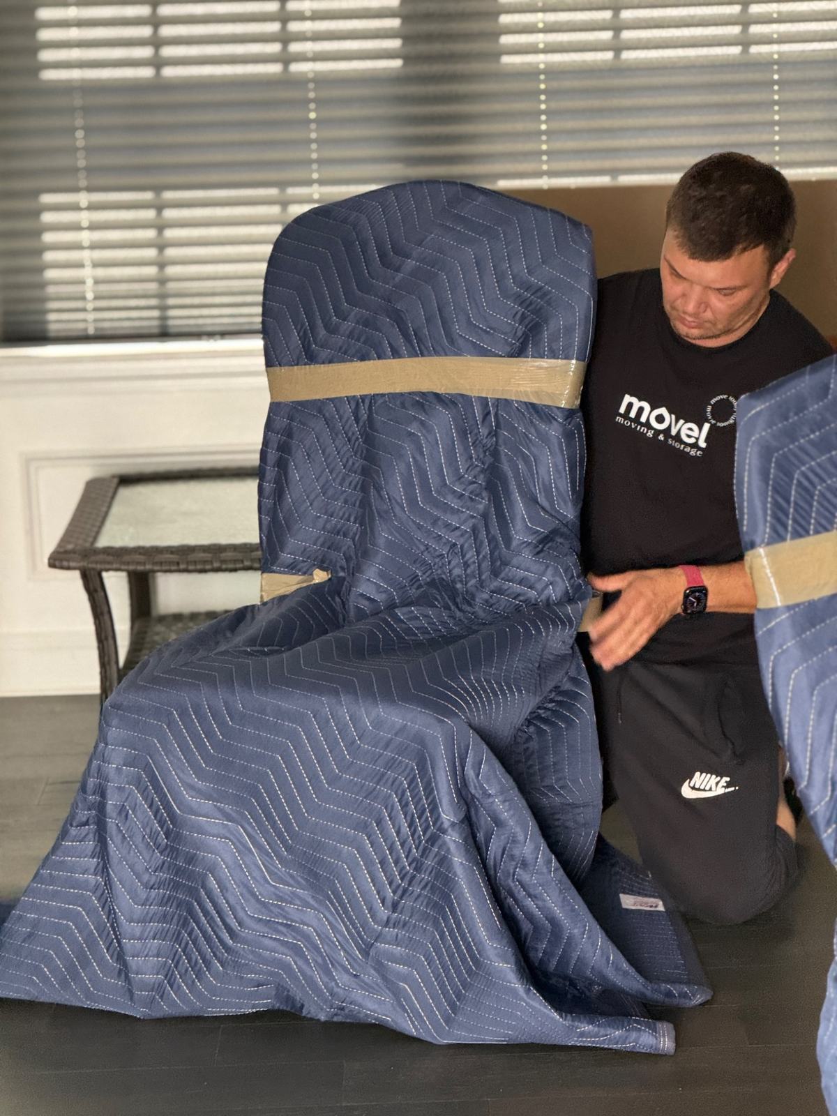 Person wrapping a chair in a blue moving blanket secured with tape indoors near a glass table.