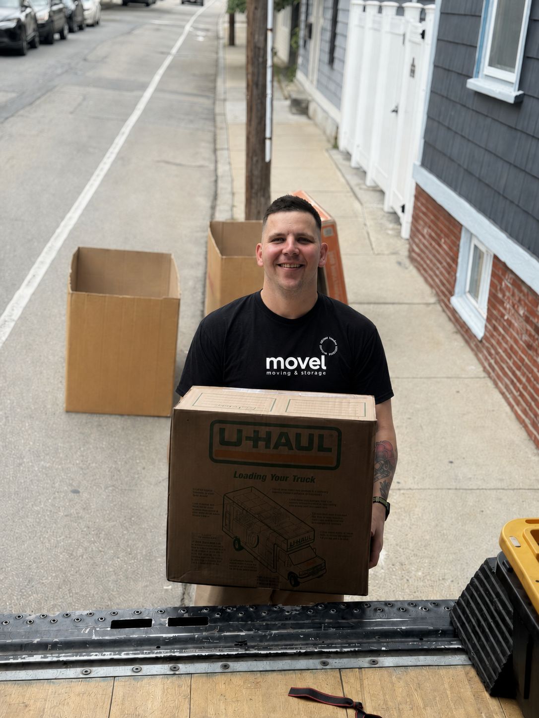 Smiling mover holding a U-Haul box on a sidewalk with empty boxes behind him.