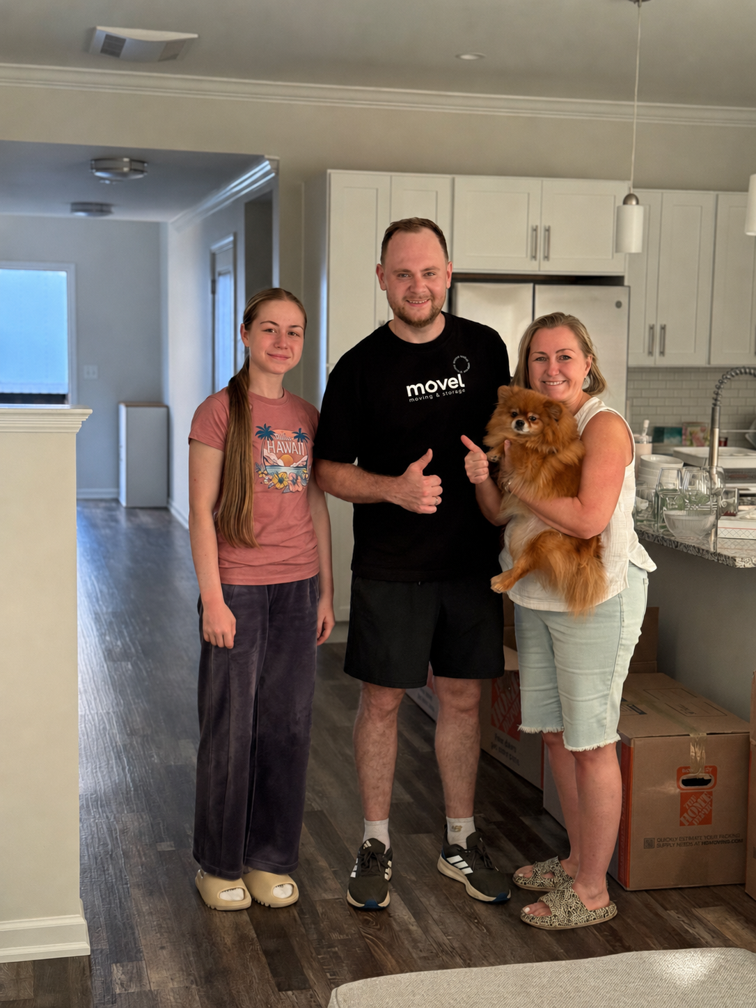 Three people smiling in a kitchen, one holding a small brown fluffy dog, with moving boxes nearby.