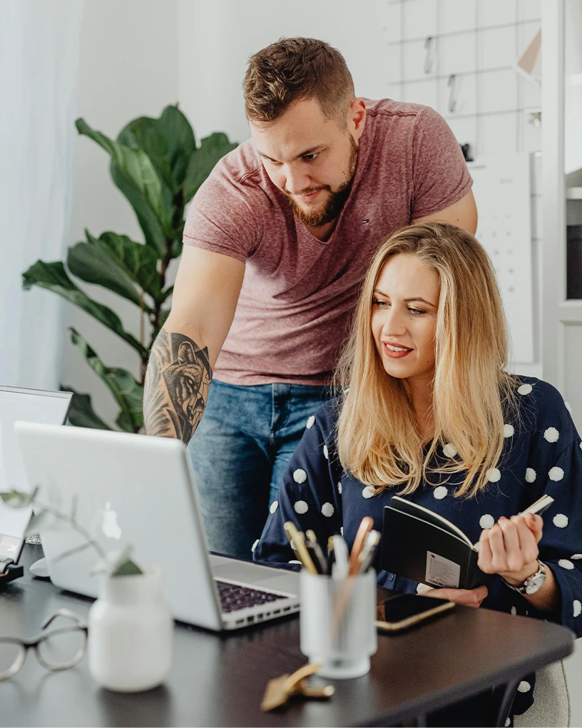 Man with tattoo on arm leaning over to help a woman with a notebook as they both look at a laptop on a desk.