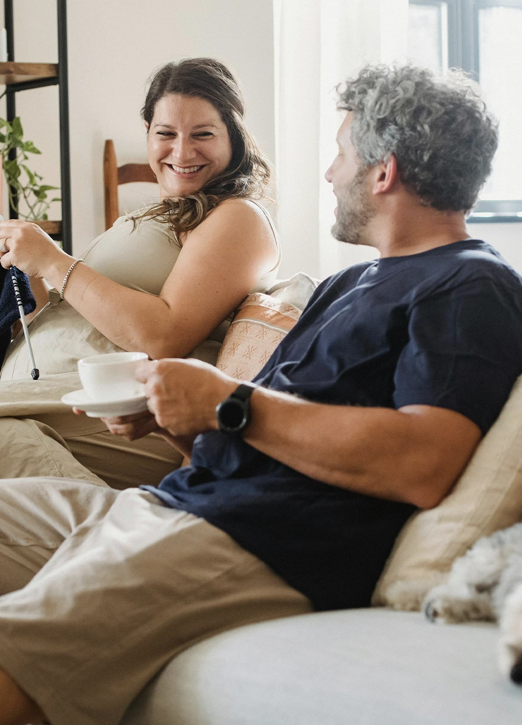Smiling couple sitting on a couch, the woman knitting and the man holding a cup and saucer, with a small dog resting beside them.