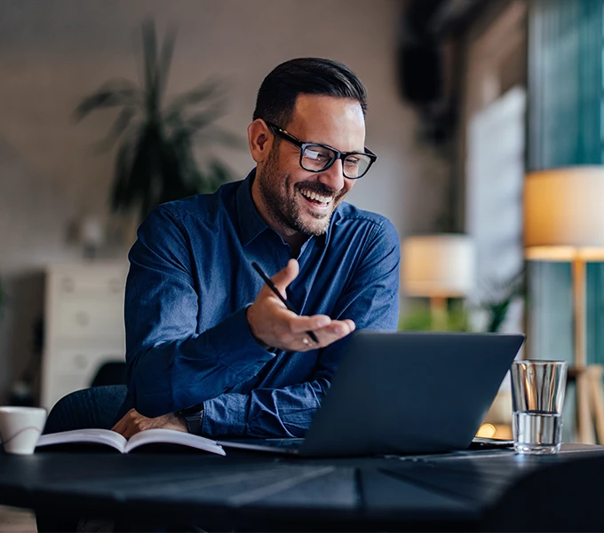 Smiling man wearing glasses and a blue shirt engaging in a video call on a laptop at a desk with a notebook and glass of water.