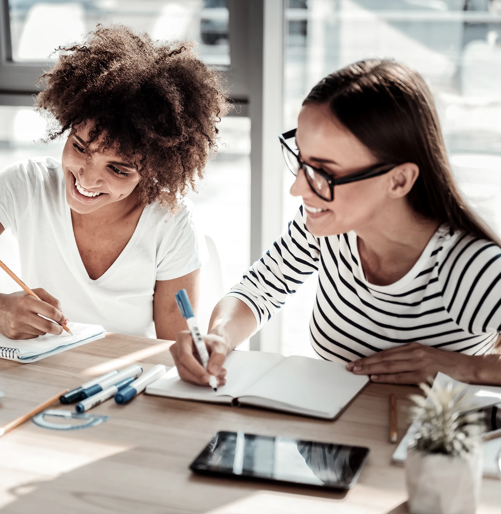 Two women smiling and writing in notebooks at a sunlit table with stationery and a tablet.