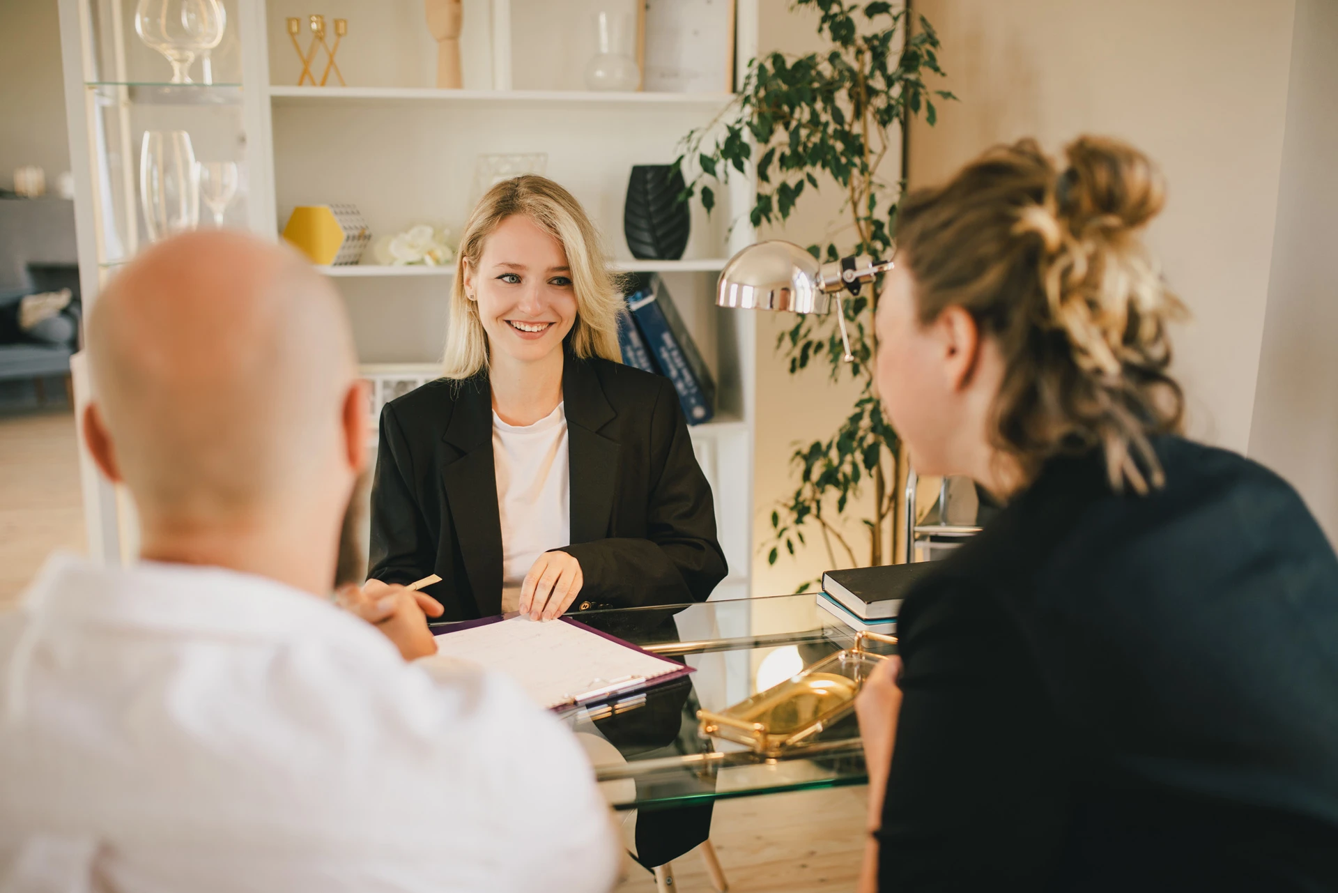 Smiling woman in a black blazer talking to two people across a glass desk in a bright office.