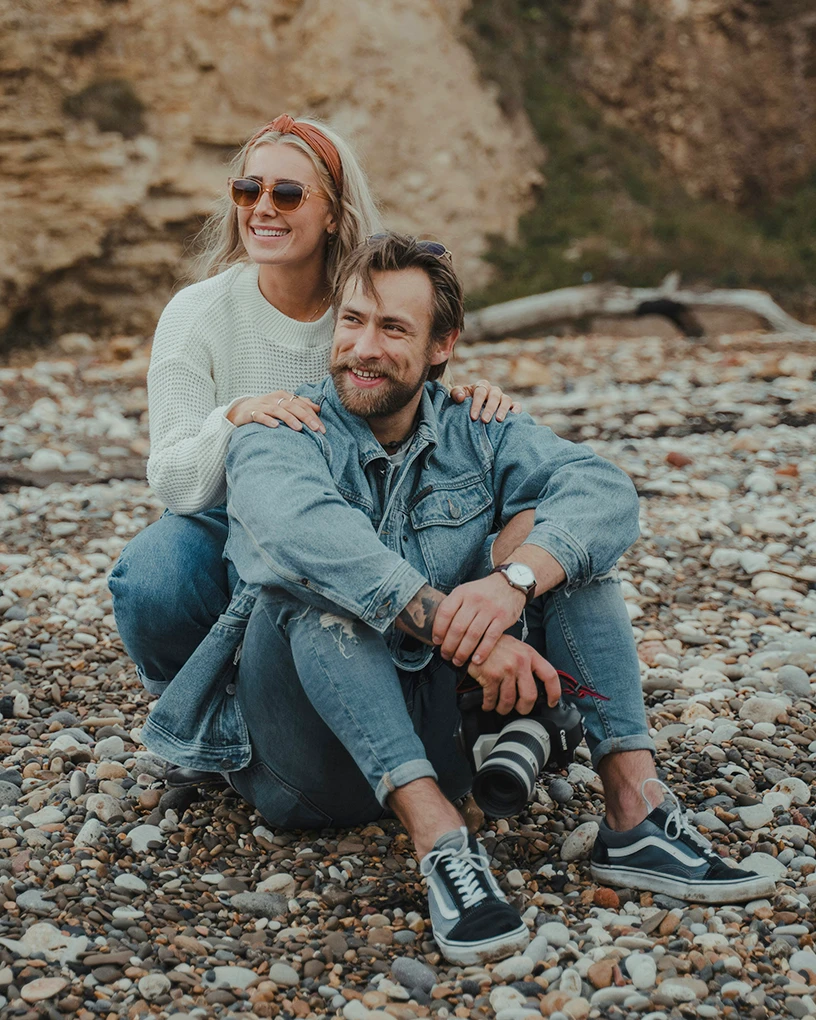 Smiling couple on a rocky beach with the man sitting holding a camera and the woman kneeling behind him with hands on his shoulders.