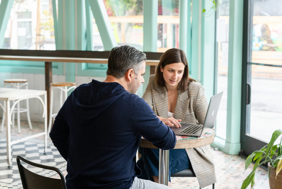 Two people sitting at a small round table in a cafe, looking at a laptop screen.