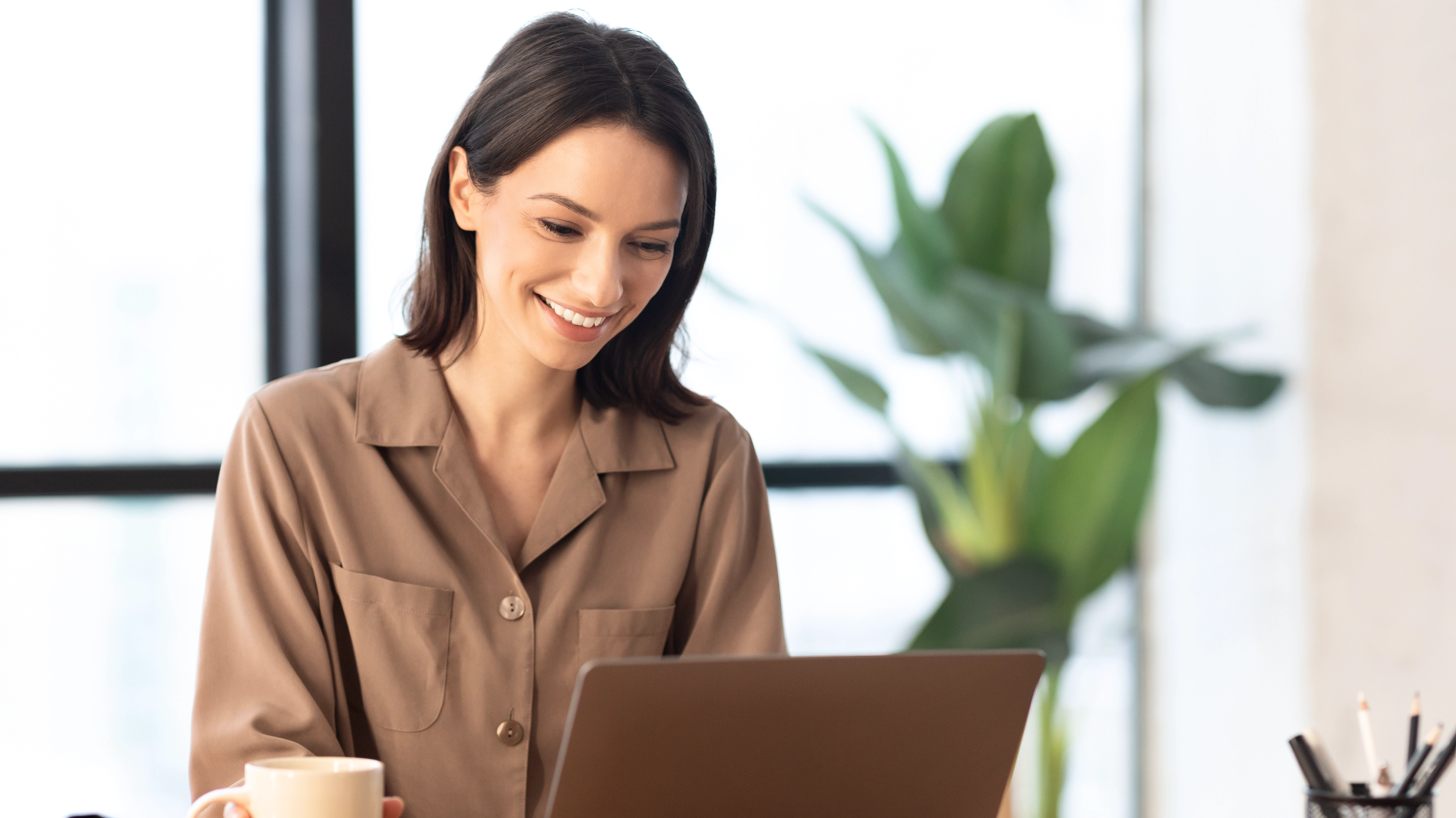 Smiling woman using a laptop at a desk with a coffee mug and a plant in the background.