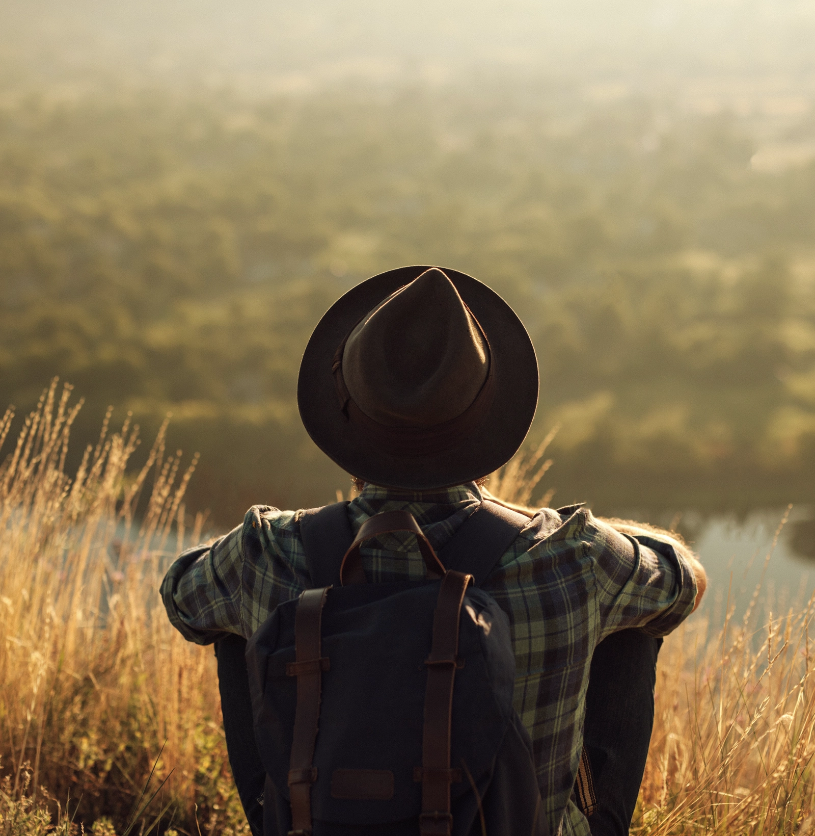 Person wearing a hat and plaid shirt sitting on a hillside overlooking a blurred landscape in warm, golden light.