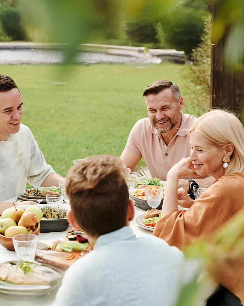 Four people enjoying an outdoor meal together at a table with food and drinks on a grassy lawn.