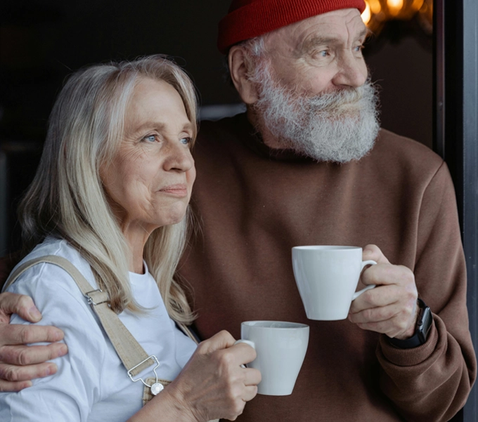 Elderly couple standing close together, each holding a white coffee cup and looking contemplatively out a window.