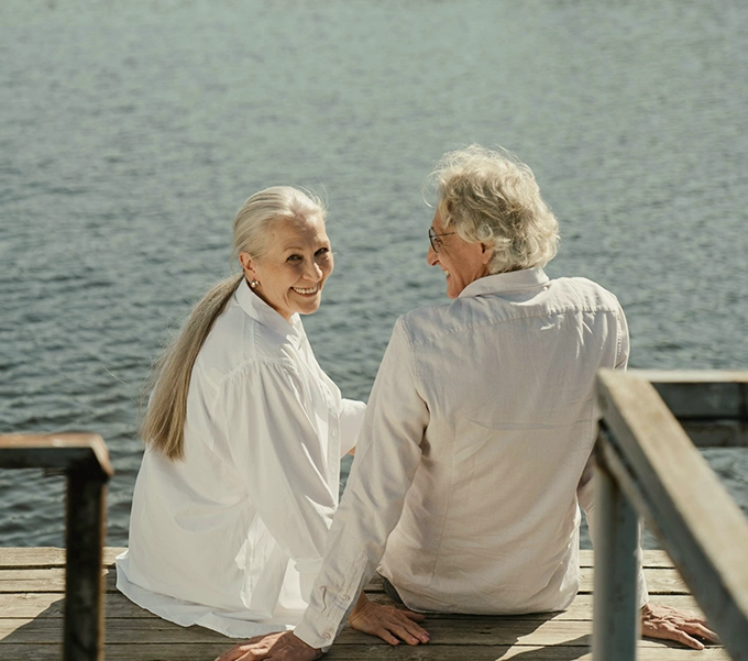 Smiling elderly couple sitting on a wooden dock by the water, holding hands and enjoying the view.