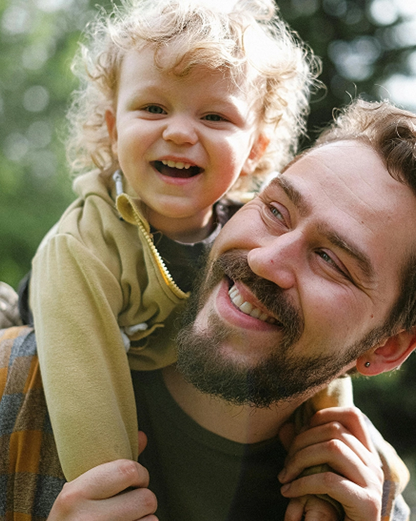 Smiling man with beard carrying a happy curly-haired child on his shoulders outdoors.