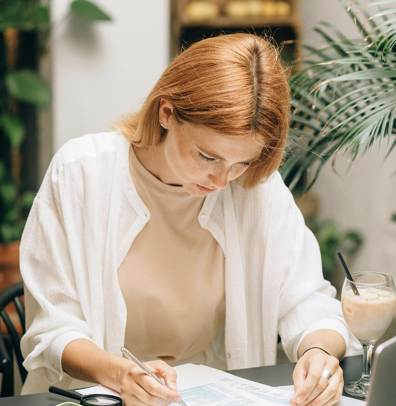 Young red-haired woman in a white shirt focused on writing on documents at a table with a magnifying glass and iced coffee.
