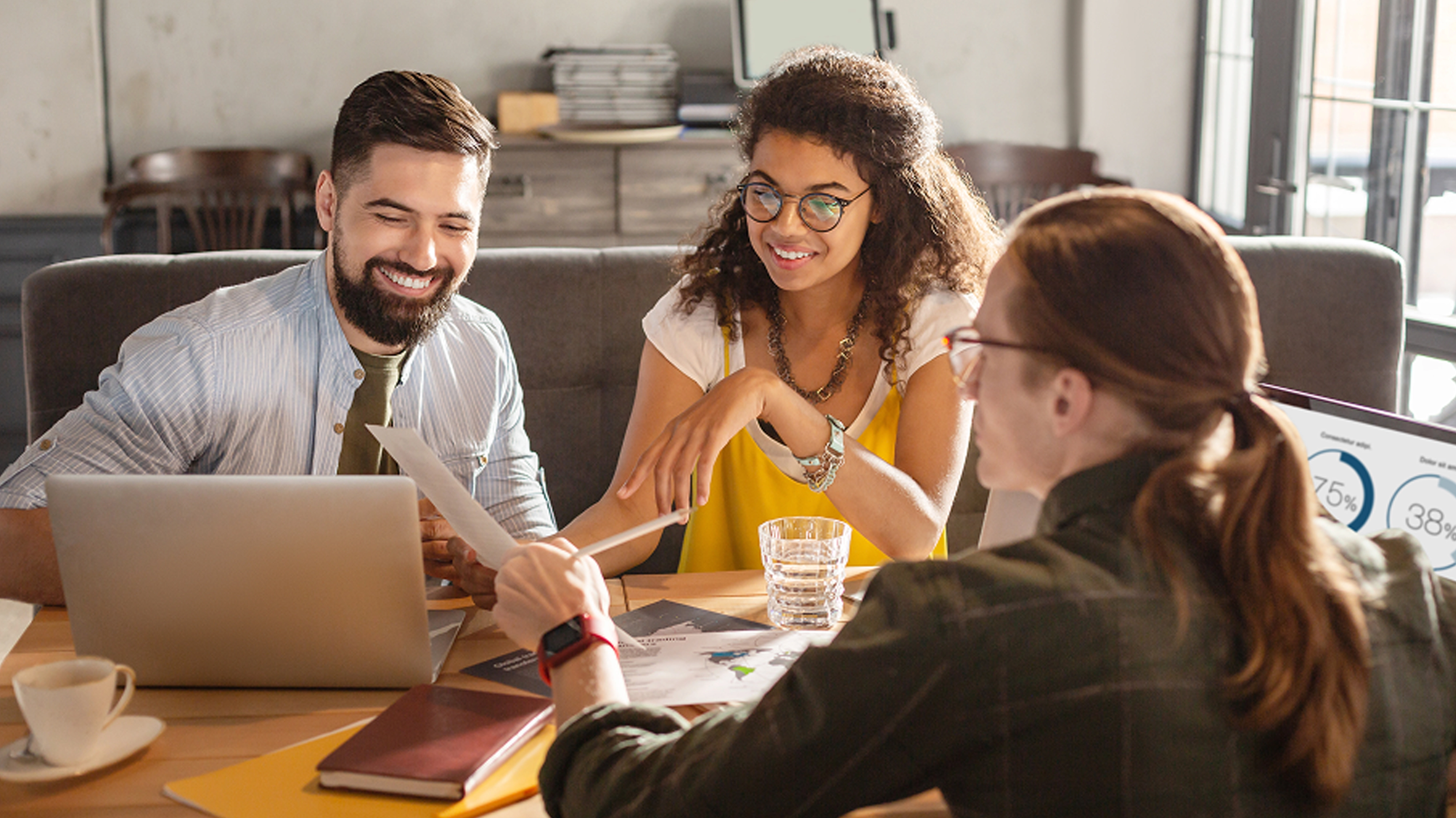 Three colleagues discussing documents around a table with laptops and charts in a casual office setting.