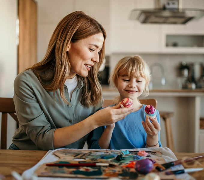 A woman and a young girl sitting at a table in a kitchen, smiling and holding painted Easter eggs.