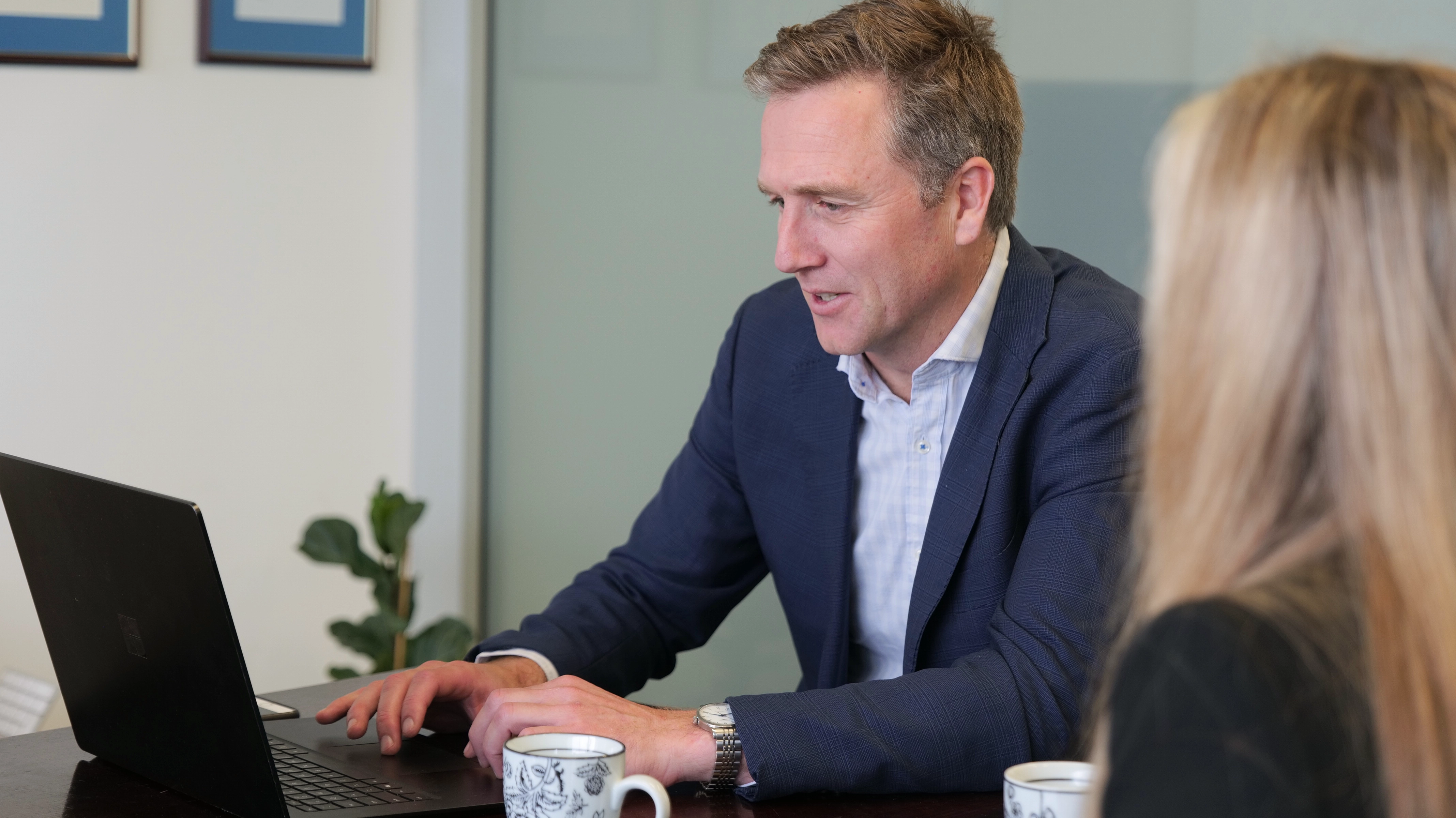 Man in blue blazer working on a laptop at a desk, speaking with a blonde woman.