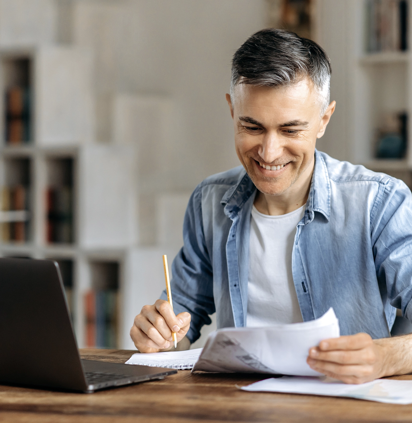 Smiling man with gray hair wearing a denim shirt, working at a wooden table with a laptop, holding papers and a pencil.