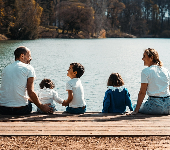 Family of two adults and three children sitting on a wooden dock by a lake surrounded by trees in autumn.