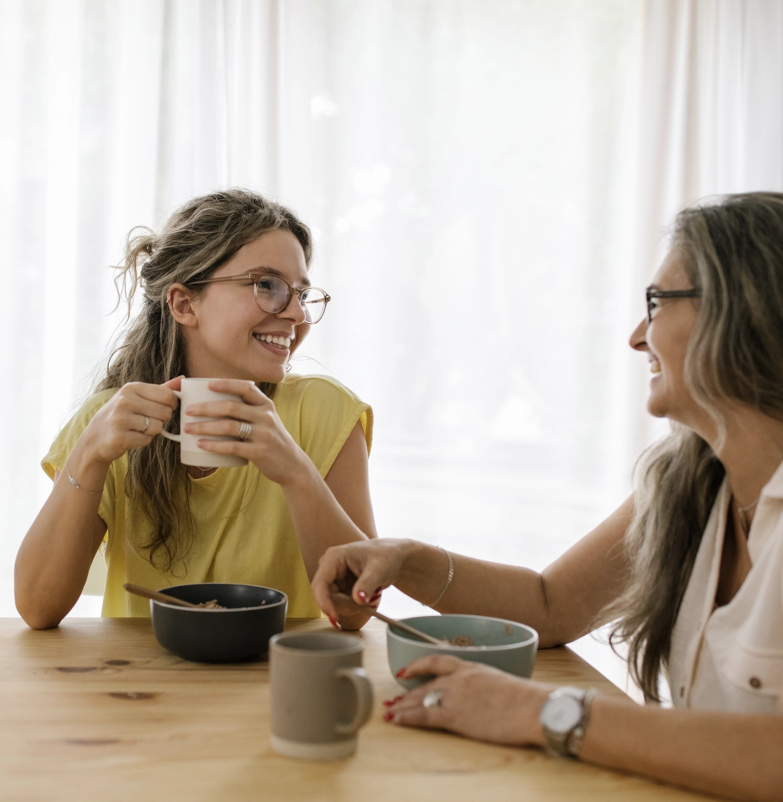 Two women smiling and chatting at a wooden table while having breakfast with bowls and mugs.