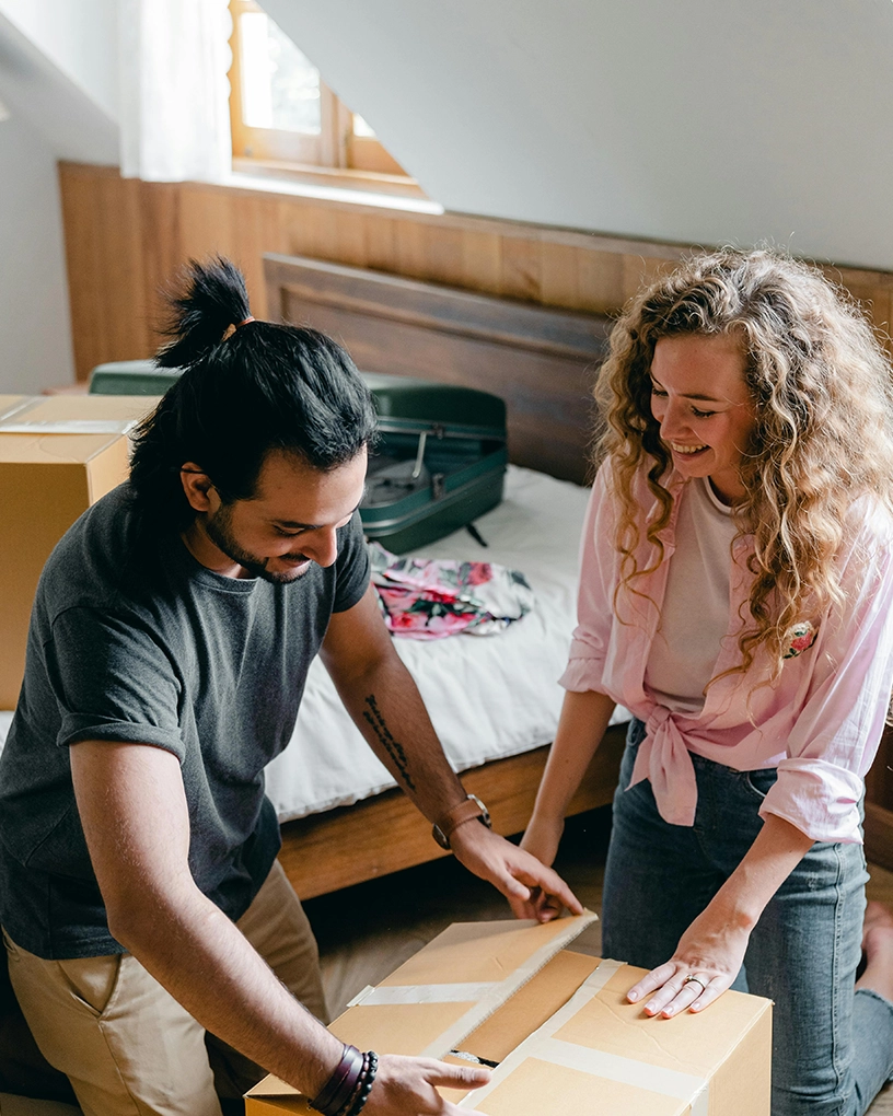 Smiling couple unpacking or packing cardboard boxes in a bedroom with a bed and suitcase in the background.