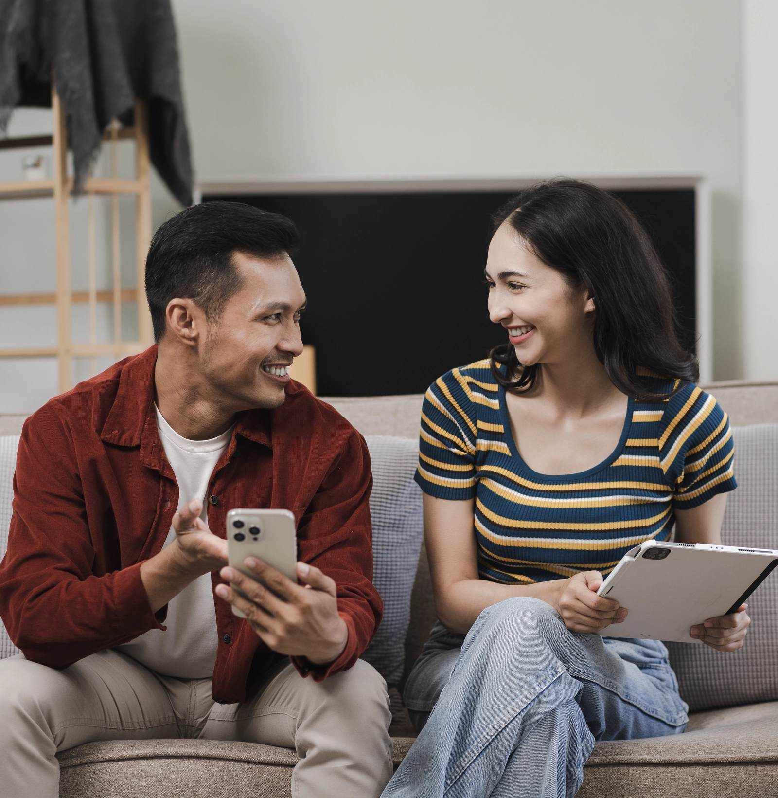 Smiling man and woman sitting on a couch, holding a smartphone and a tablet while looking at each other.