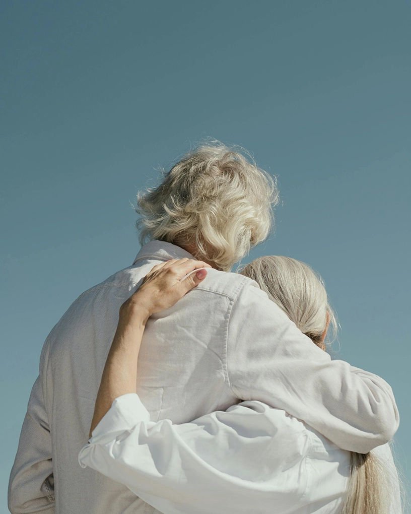 Rear view of an elderly couple embracing outdoors against a clear blue sky.