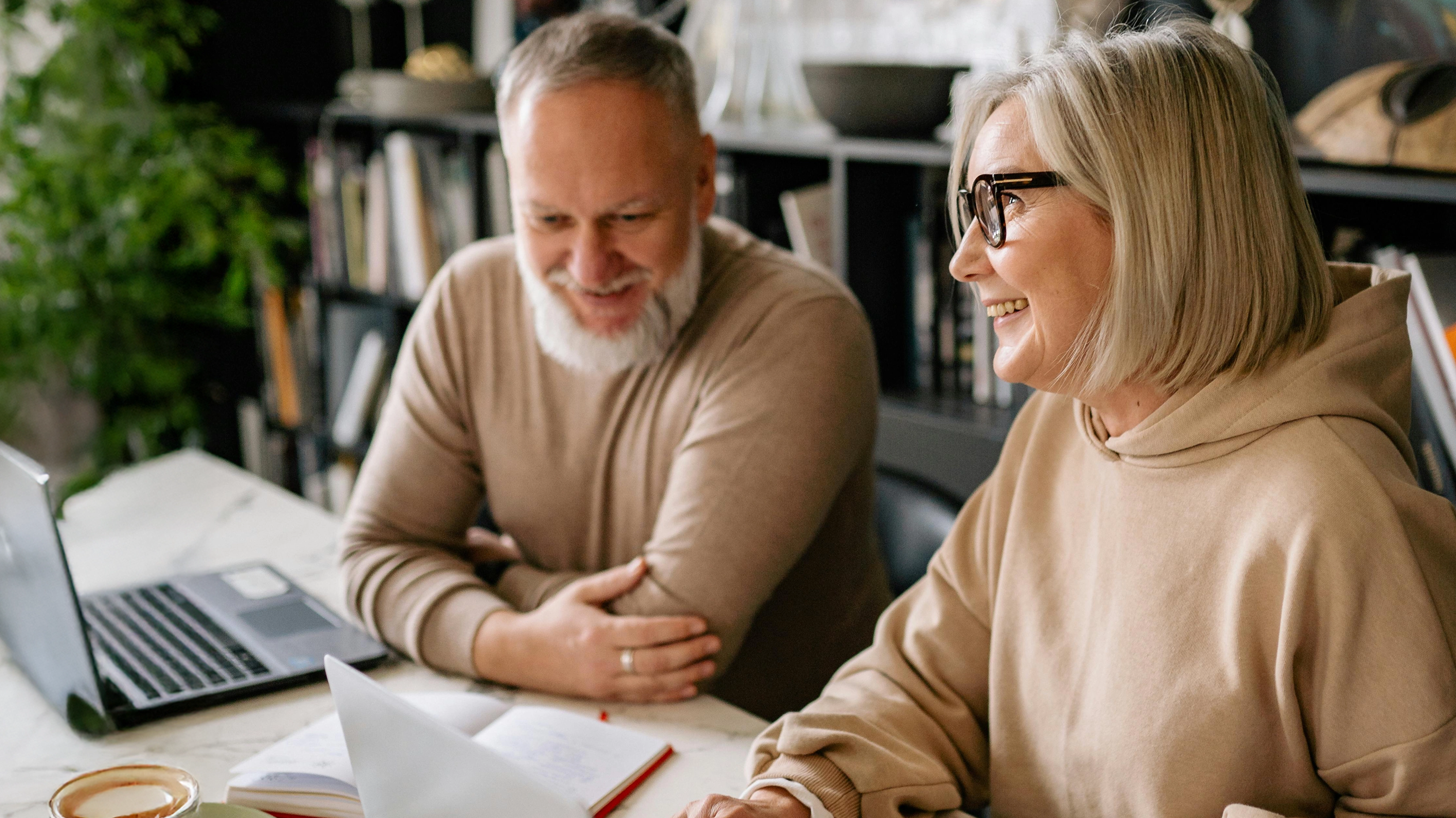 Smiling mature man and woman sitting at a desk with laptops and notebooks, engaged in a friendly discussion.