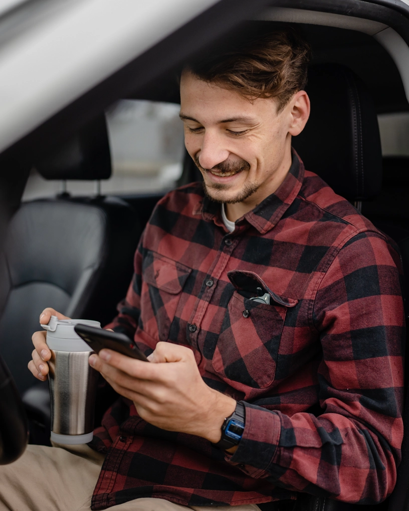 Smiling man in a red and black plaid shirt sitting in a car, holding a travel mug and looking at his smartphone.