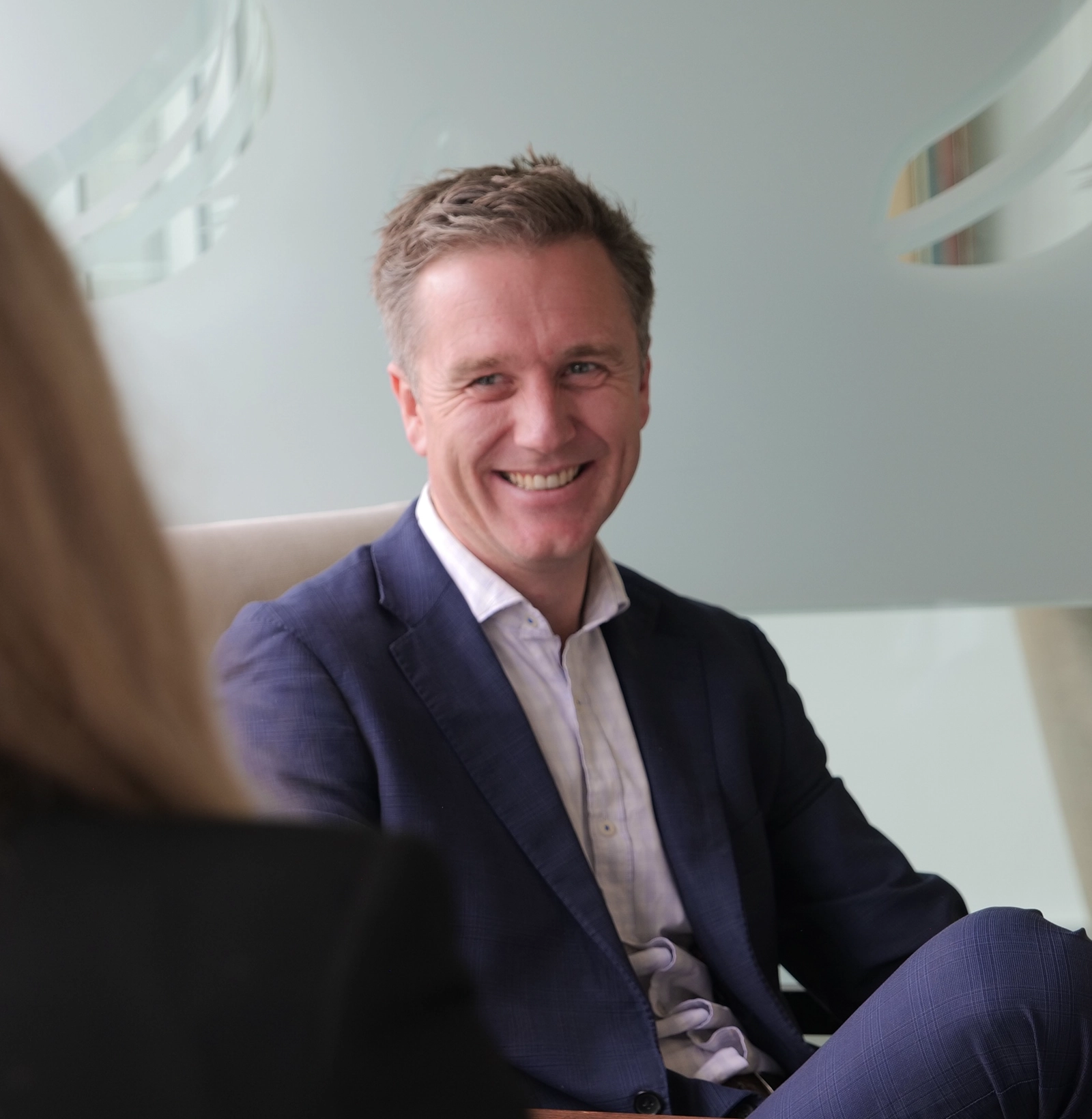 Smiling man in a blue suit sitting and talking to a woman in an office setting.