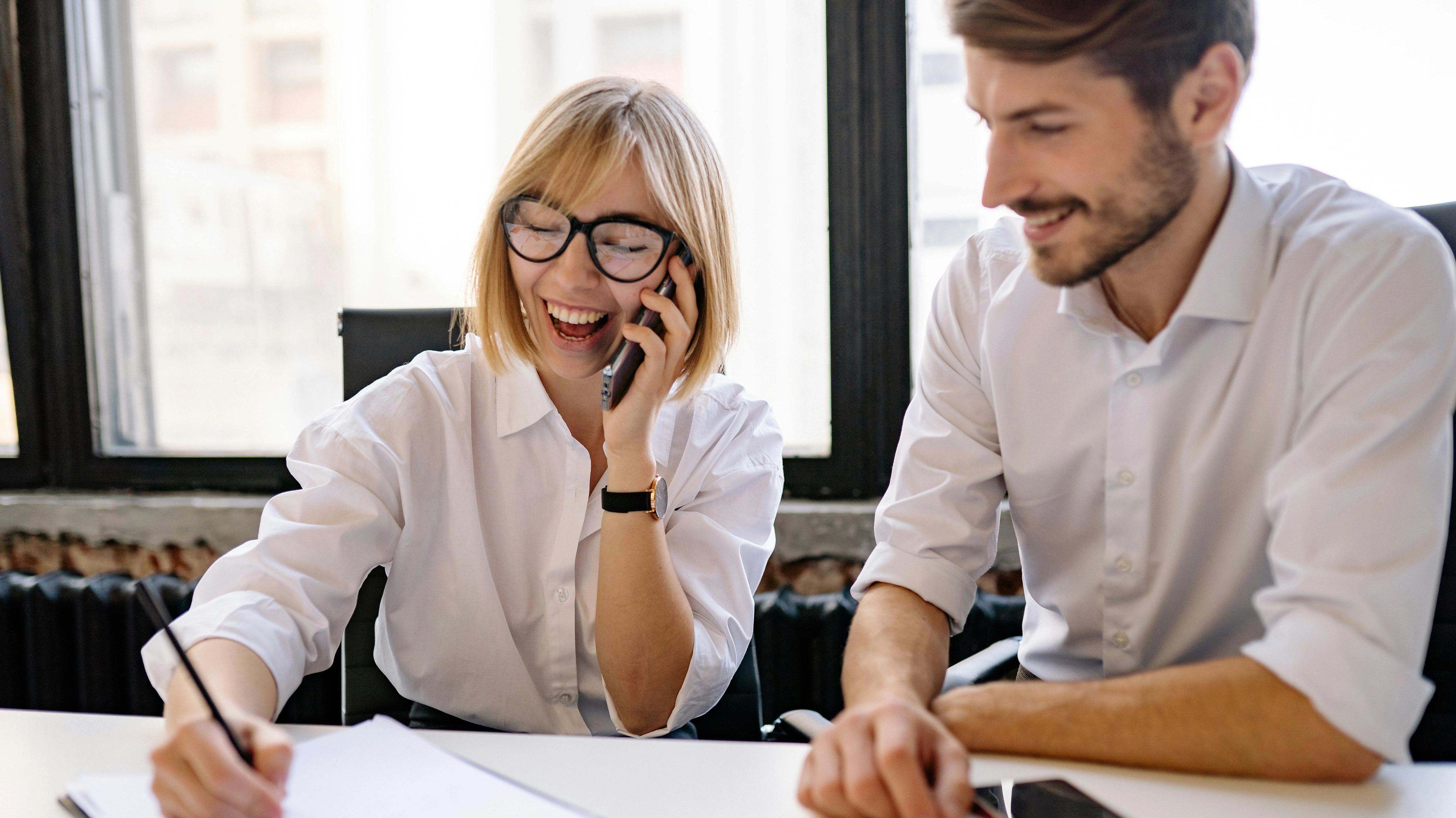 Two young professionals in white shirts collaborating at a desk while the woman talks on a phone and laughs.