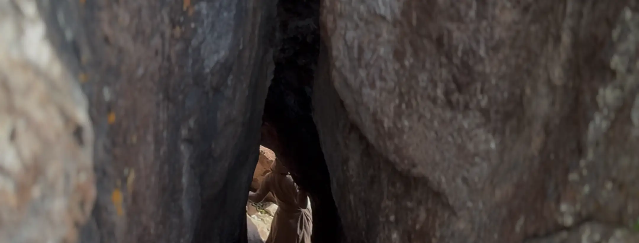 Person in a brown coat standing between large dark rock formations, viewed through a narrow gap.