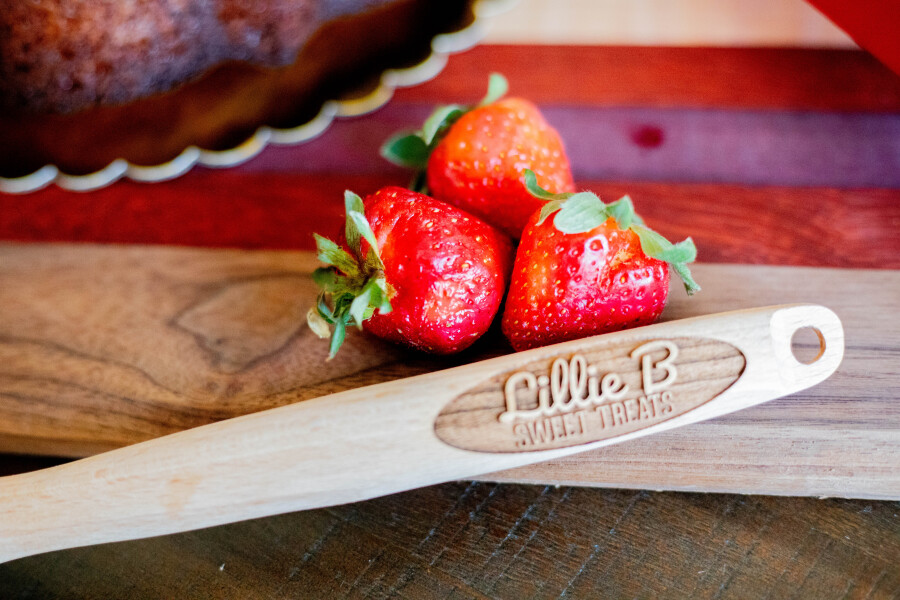 Three fresh strawberries on a wooden board with a wooden spatula engraved 'Lillie B Sweet Treats'.