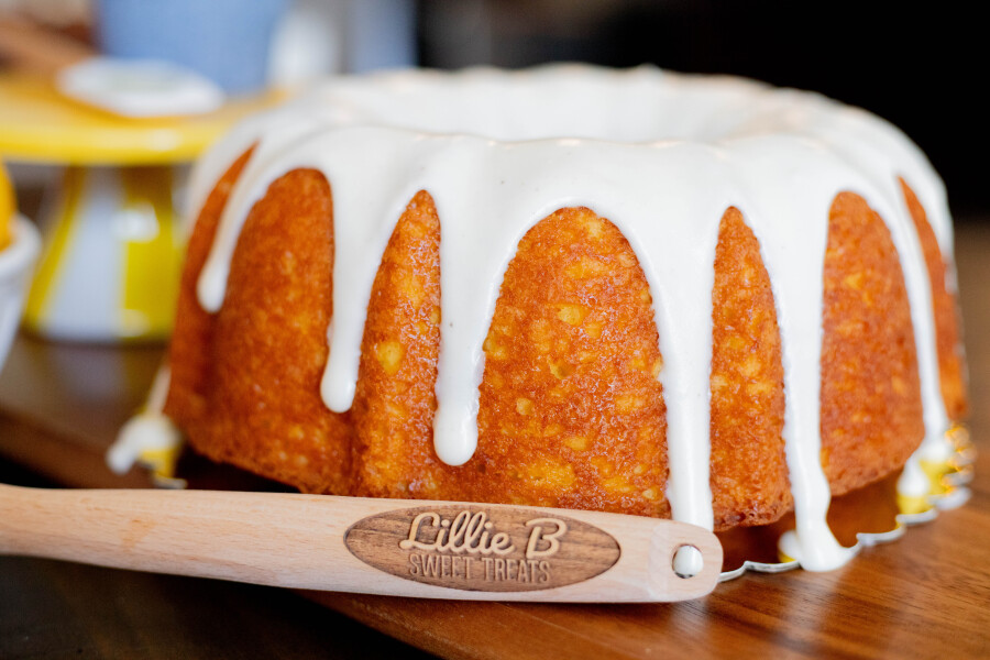 Bundt cake with white icing dripping down sides on wooden table with wooden spatula reading Lillie B Sweet Treats.