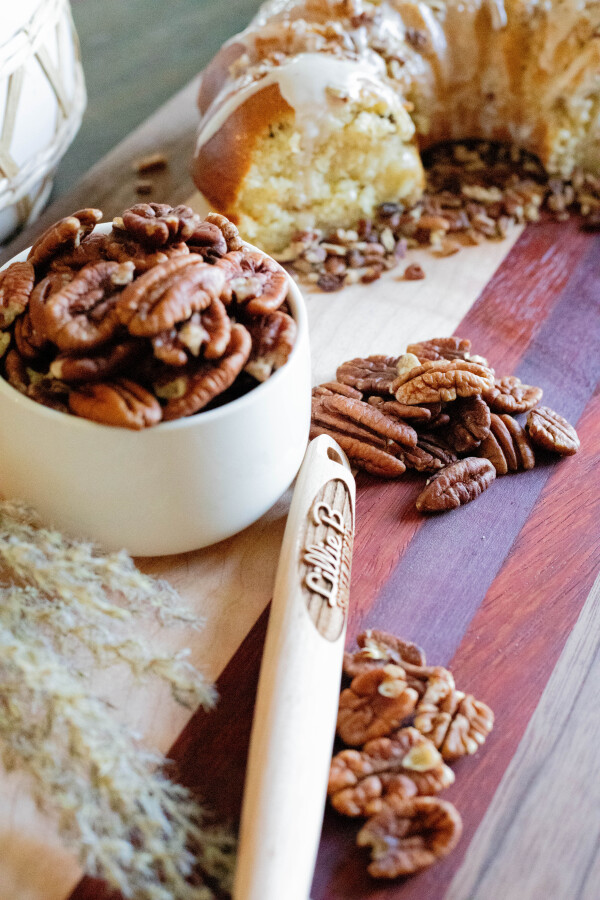 Pecans in a white bowl and scattered on a wooden board near a bundt cake and wooden spoon.