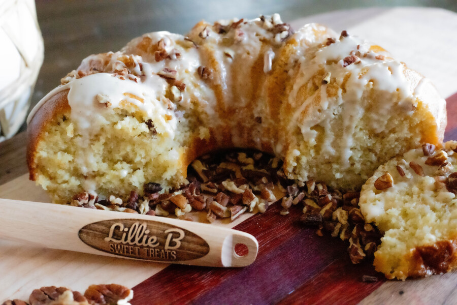 Bundt cake with white icing and chopped pecans on a wooden board next to a wooden spatula.