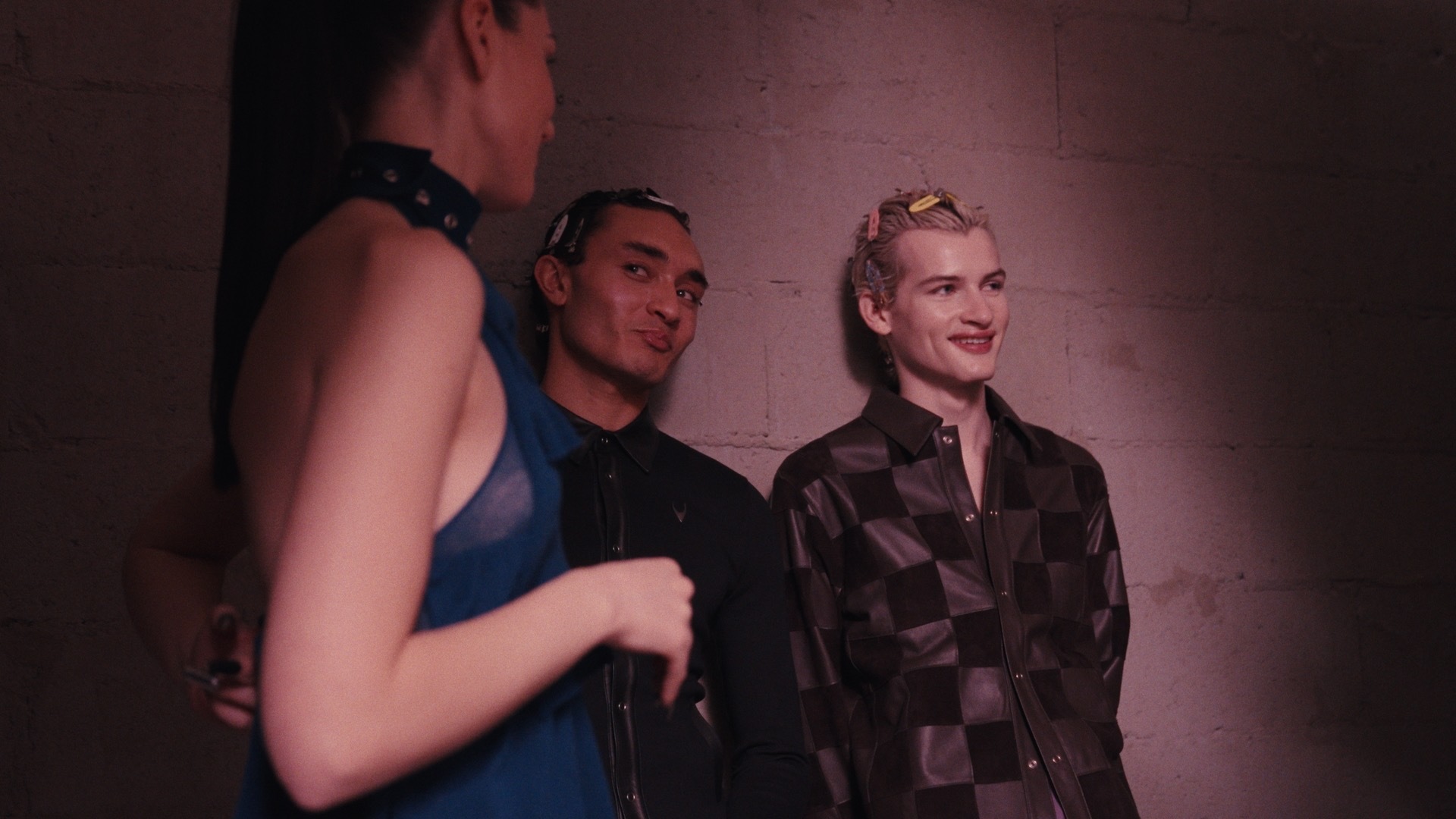 Three people standing against a concrete wall in dim purple lighting