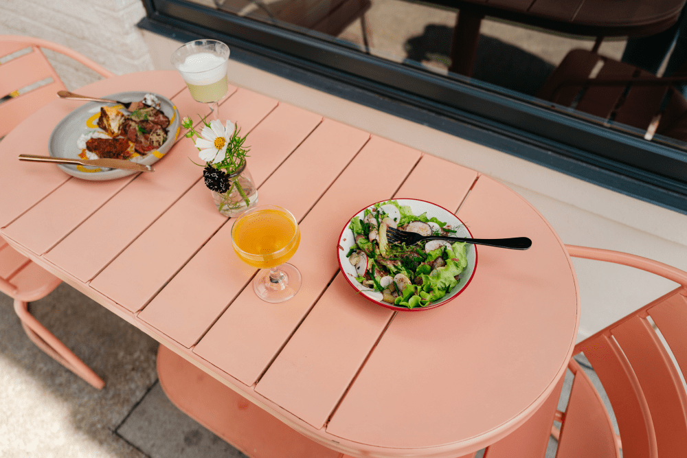 Outdoor café table with a salad, plated entrée, orange cocktail, and a small vase of flowers beside a window.
