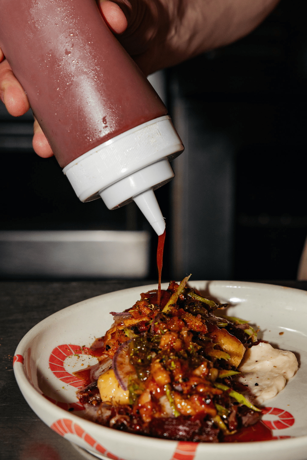 Close-up of a hand squeezing reddish sauce from a plastic bottle onto a plated dish
